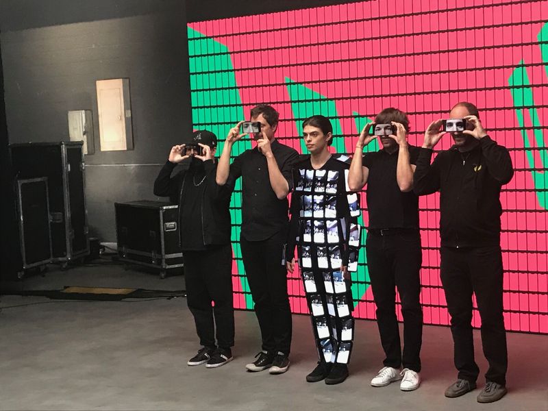 Five crew members in black holding phones up to their eyes, and one in a suit covered in phones, in front of a wall of phones showing a red and green pattern.