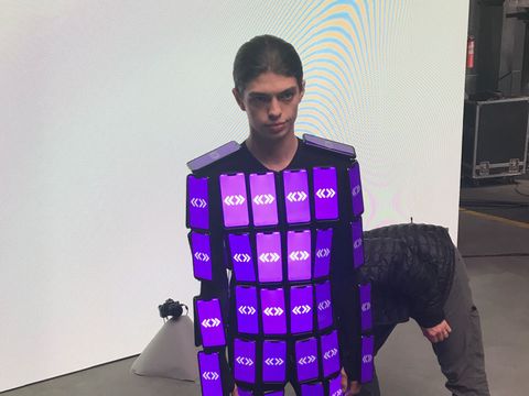 A dancer standing in a suit covered in phones, all showing the same purple pattern.